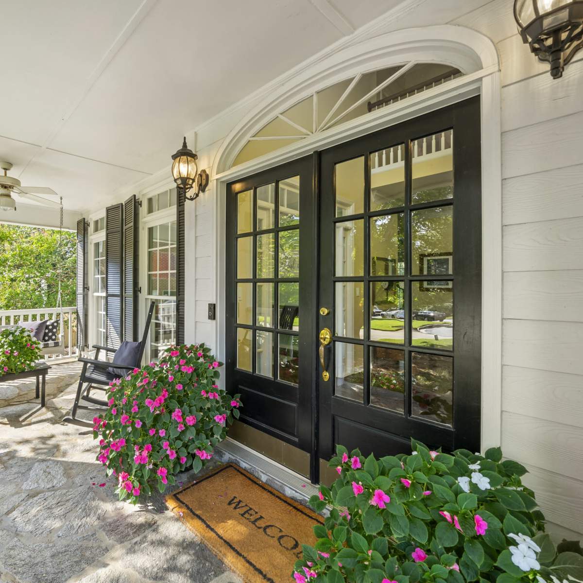 A front porch with rocking chairs, potted flowers, and a welcome mat in front of large double doors.