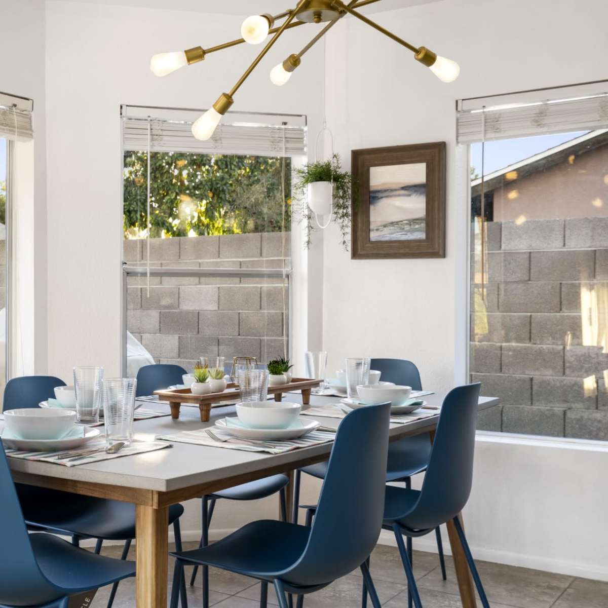 A modern dining area features a wooden table set for eight with blue chairs, surrounded by large windows showcasing a landscaped backyard.