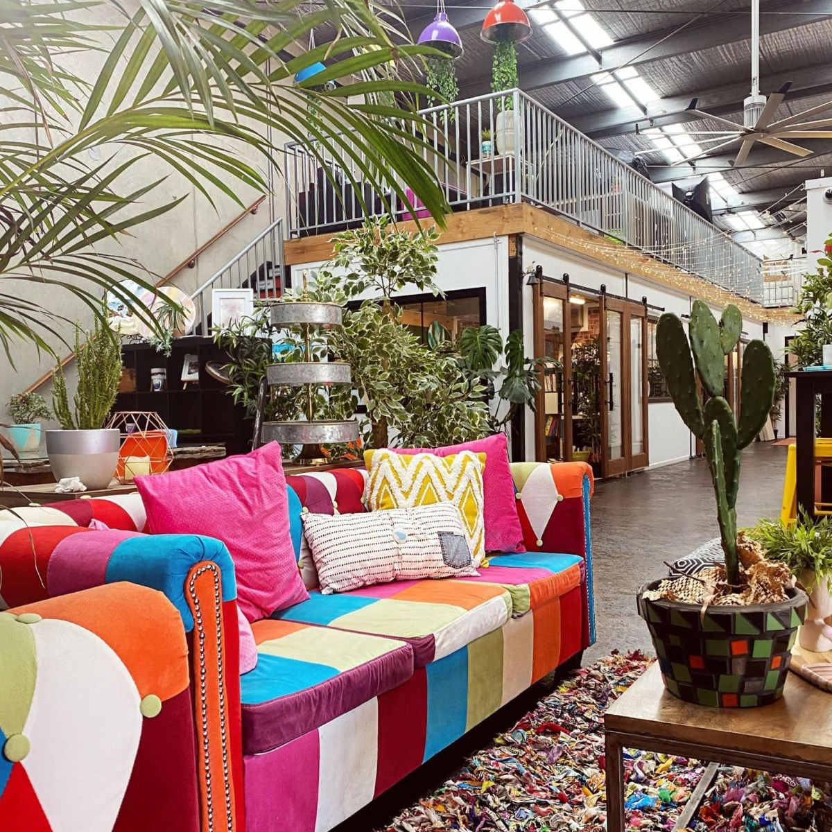 A brightly colored patchwork sofa sits in a lush, plant-filled indoor space with an upper level and decorative lights.