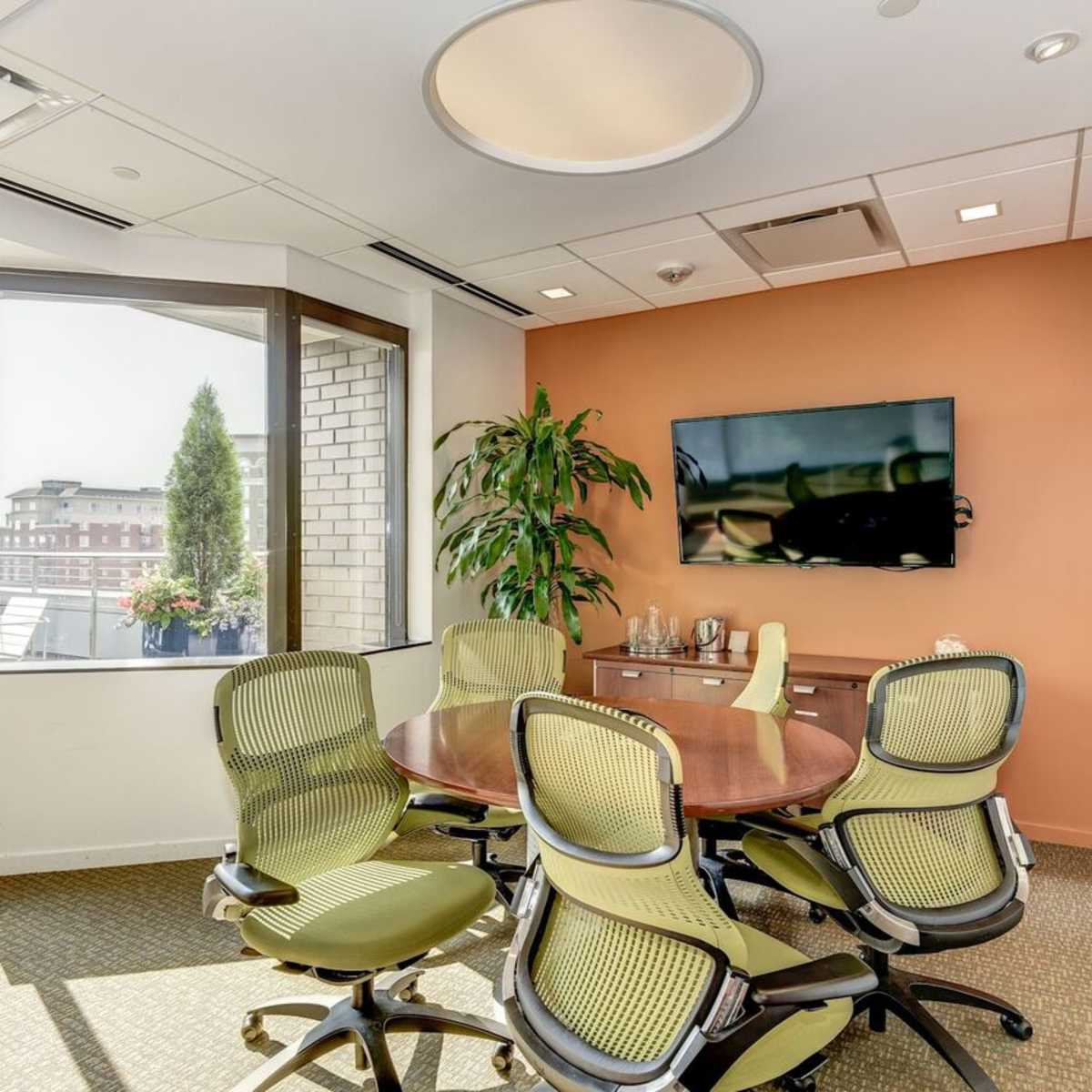 A corner conference room features a round table surrounded by green chairs, with large windows offering a view and a television mounted on the wall.