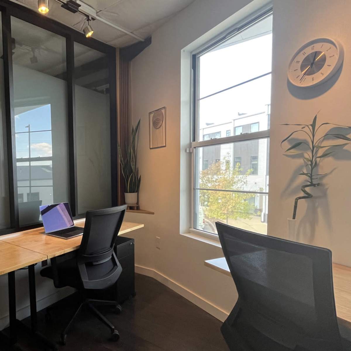 A modern office space with two desks, each equipped with a laptop, against a backdrop of large windows and partitioned glass walls.