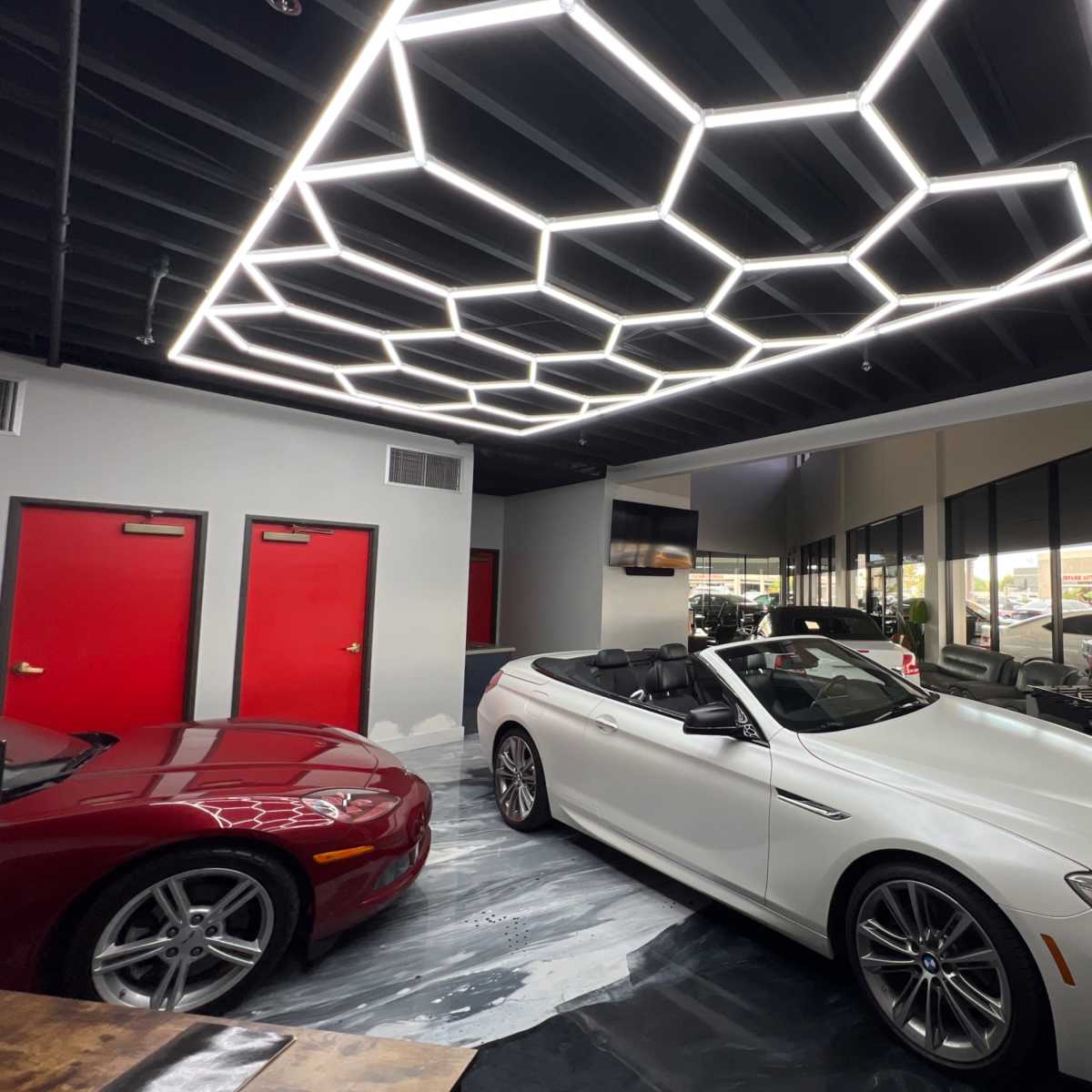 A sleek white convertible car parked in a modern showroom featuring a honeycomb-patterned light fixture and red doors.