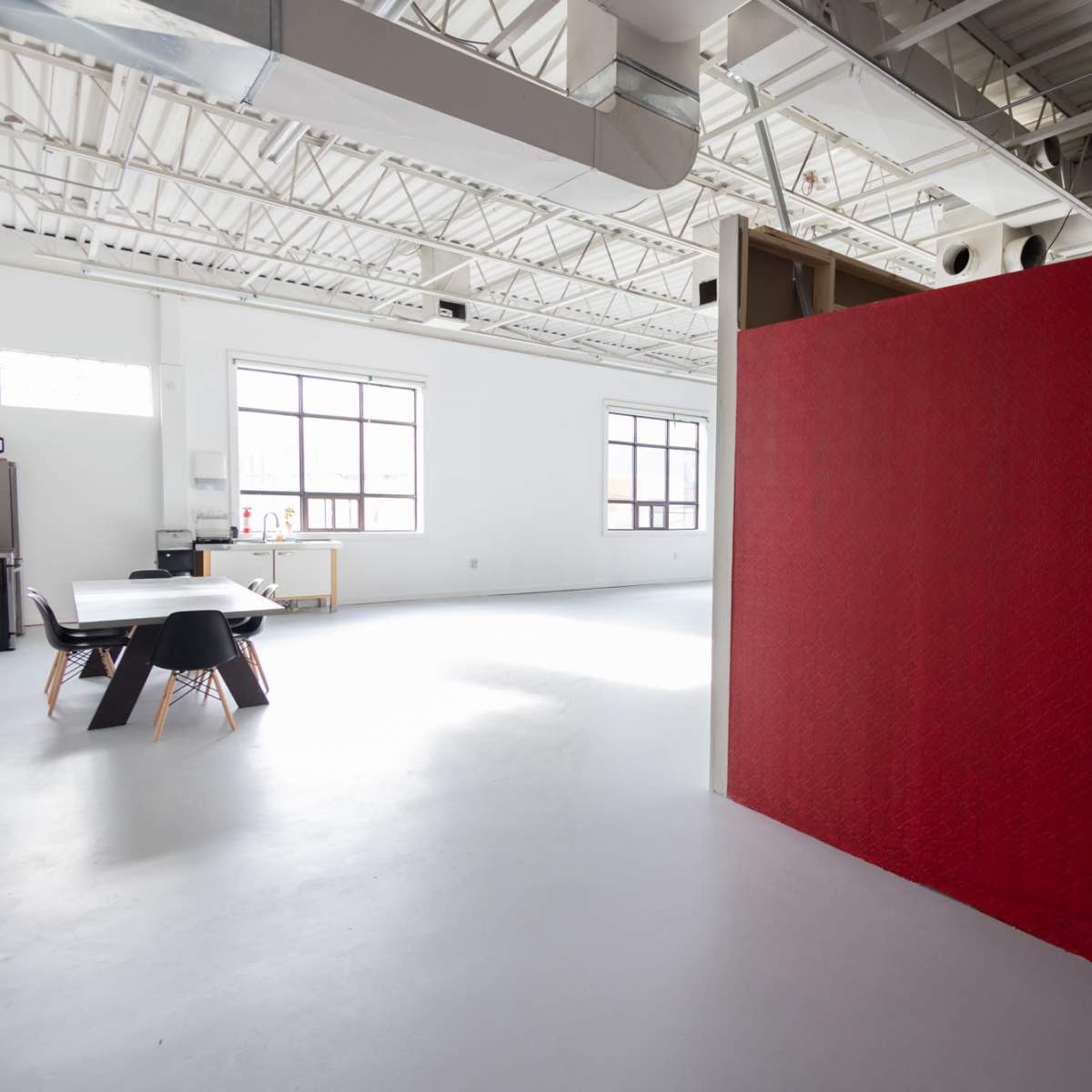 A large, bright room with high ceilings features a red partition and a table with chairs in the background.