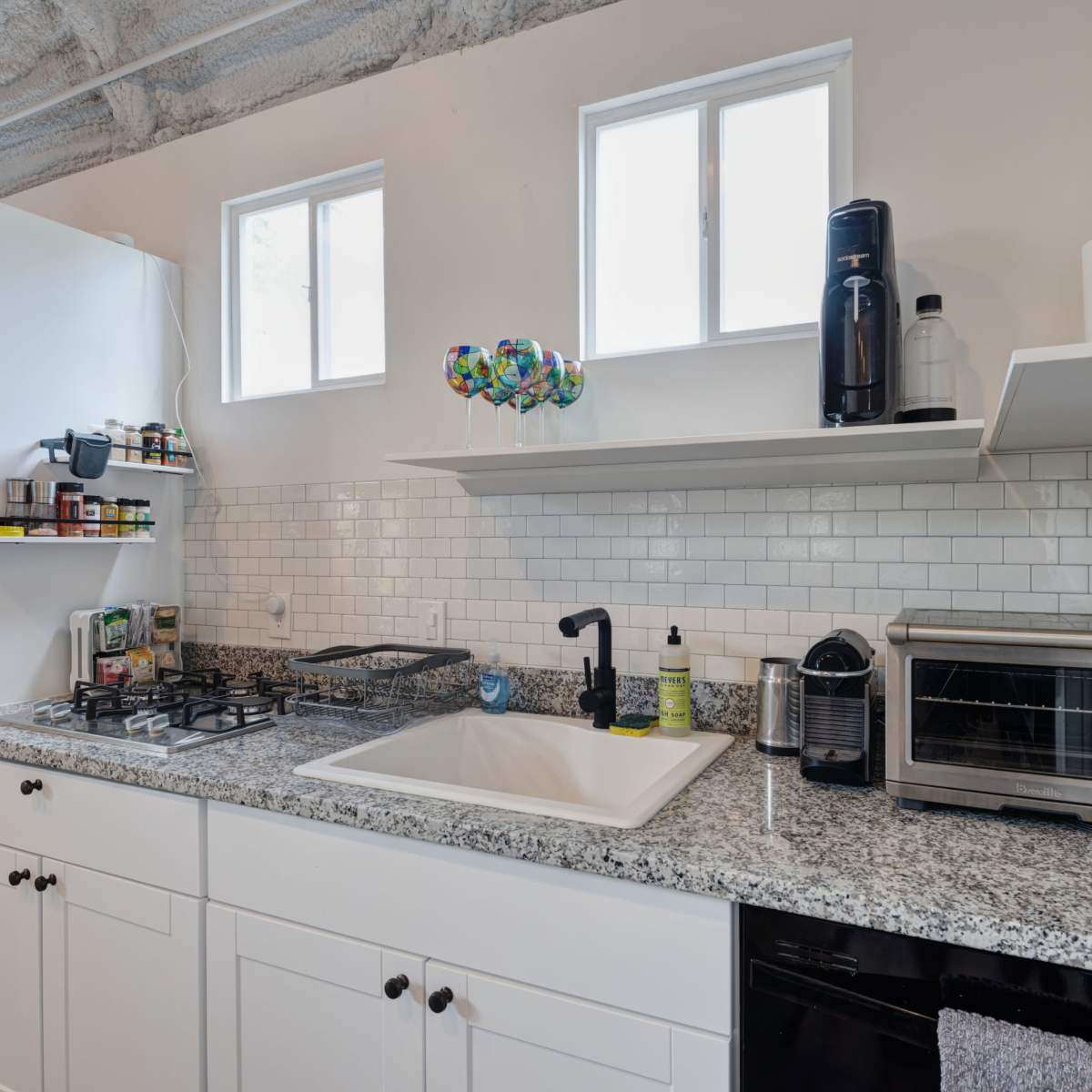 A modern kitchen features a granite countertop with a sink, gas stove, microwave, and shelves holding various kitchen items and decor.