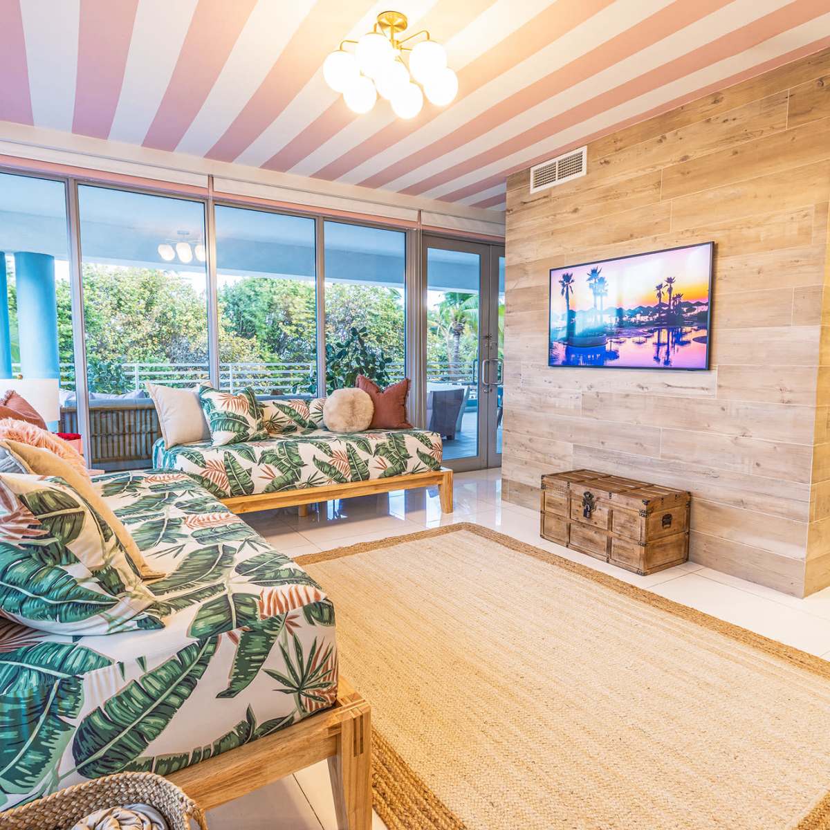 The room features two leafy-patterned daybeds, a wooden television stand, and large windows with views of greenery, all under a striped ceiling.