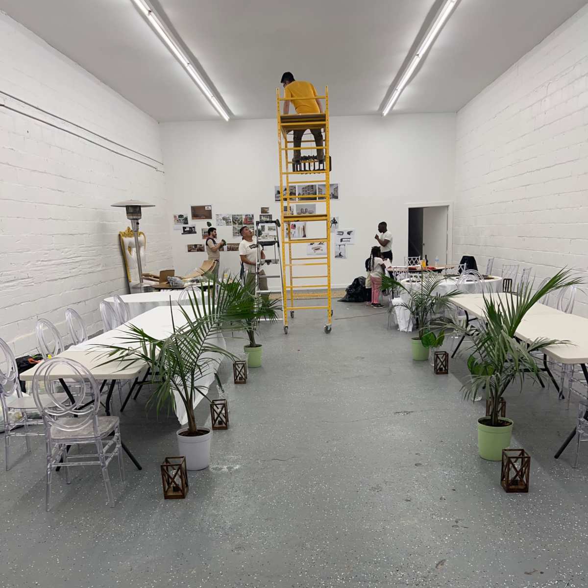 A spacious room features two long tables lined with clear chairs, potted plants on the floor, and a tall ladder against the wall where a person is working.