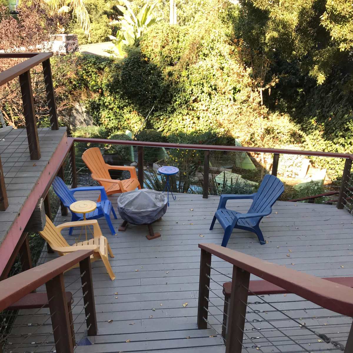 A wooden deck with brightly colored chairs surrounds a small table and fire pit, overlooking a lush, green garden.