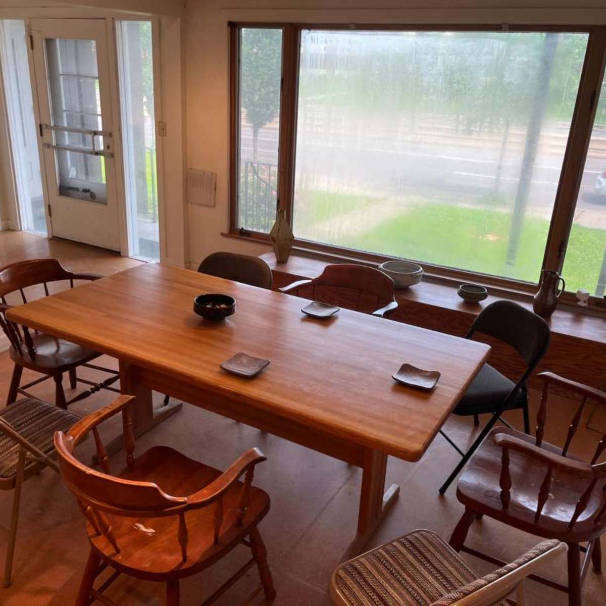 A wooden dining table surrounded by eight chairs is positioned in a room with large windows, revealing a view of the outside.