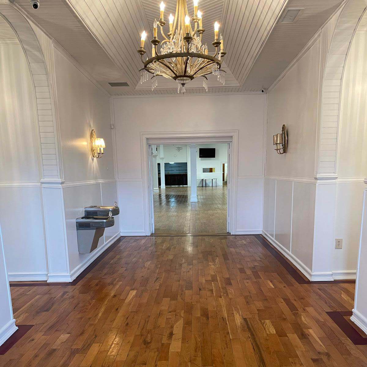 A well-lit hallway with polished wooden floors leads to an open room at the far end, featuring a chandelier and wall sconces.