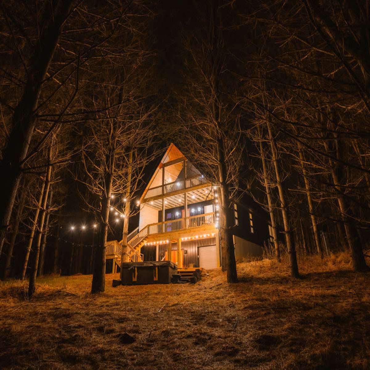A wooden A-frame house is illuminated by string lights and stands among tall, bare trees at night.