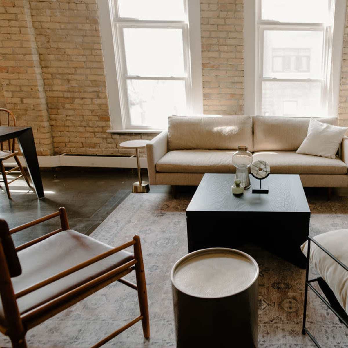 A bright living room with large windows, featuring a beige sofa, a black coffee table, a floor lamp, and a patterned area rug.
