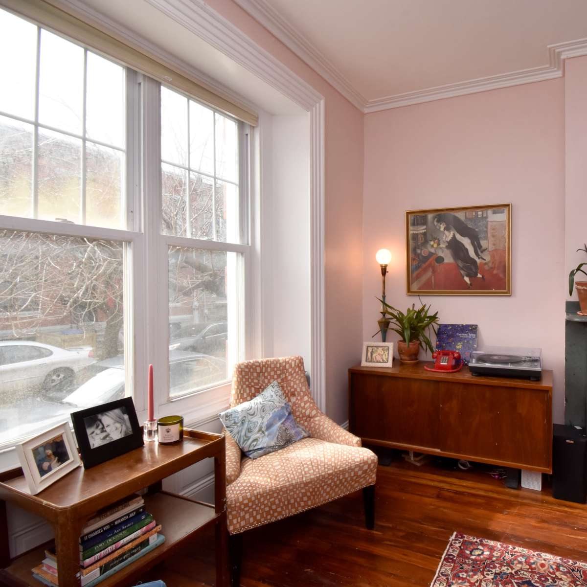 A cozy corner of a living room features a patterned chair next to a wooden table, framed by large windows and a decorative fireplace with plants and artwork.