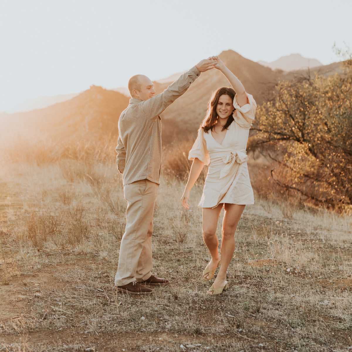 A couple dances hand-in-hand on a grassy hillside with mountains in the background during sunset.