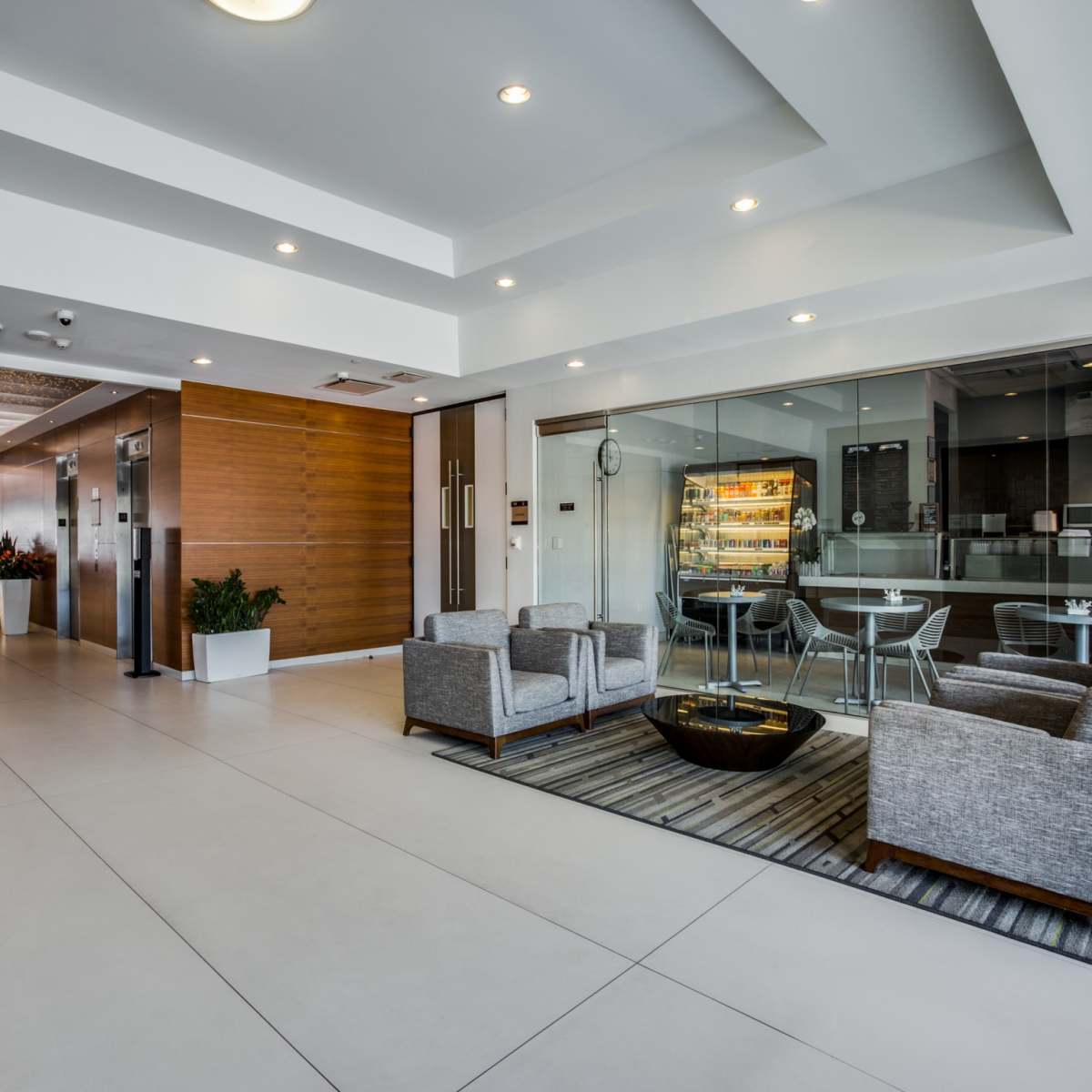A modern lobby with two grey seating areas, a circular coffee table, and a glass-walled meeting space in the background.
