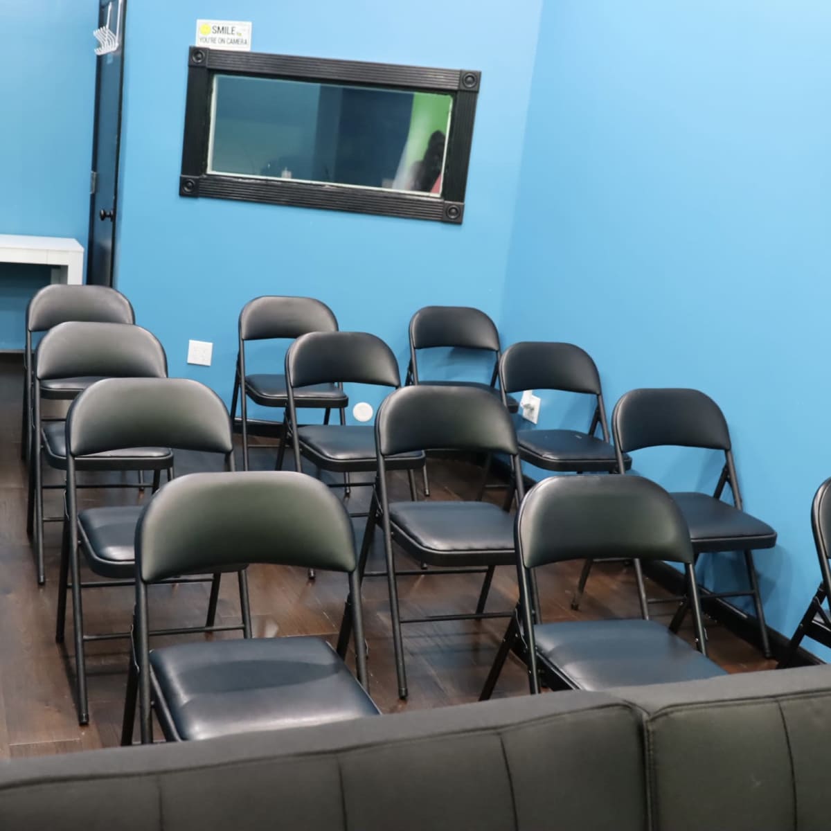 A small, blue-walled room with several black folding chairs arranged in rows facing a wall mirror.