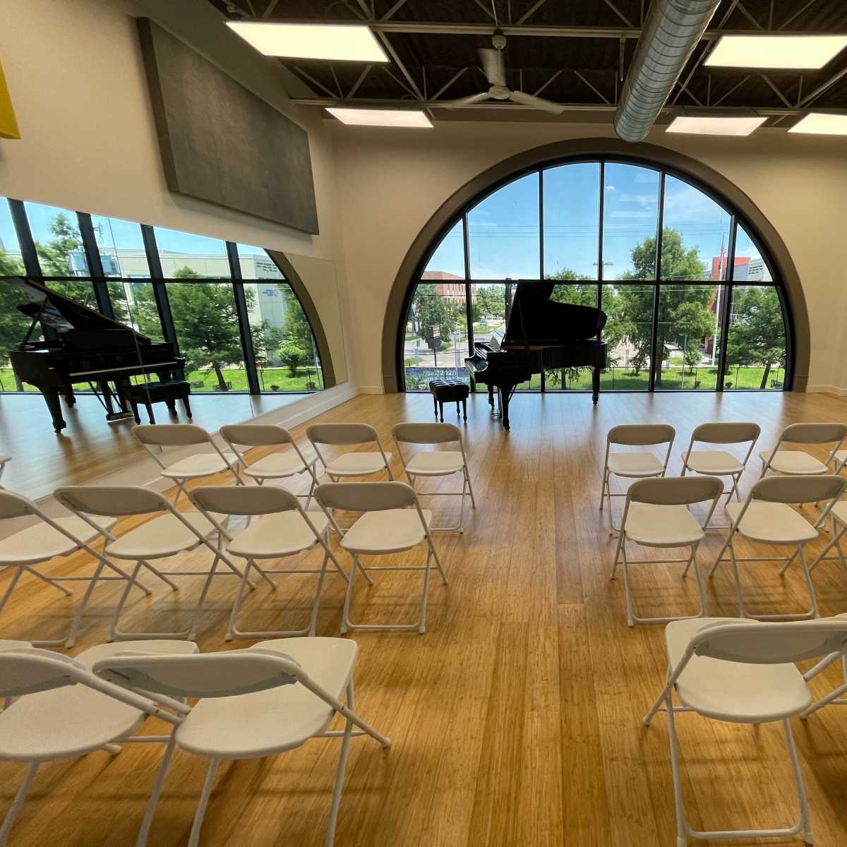 A spacious music room with two grand pianos and white folding chairs arranged in front of them, set against large windows that overlook a green landscape.