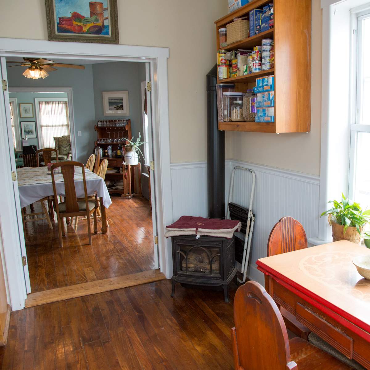 A bright kitchen with a red table and wooden chairs, opening into a dining area with a table set for a meal.