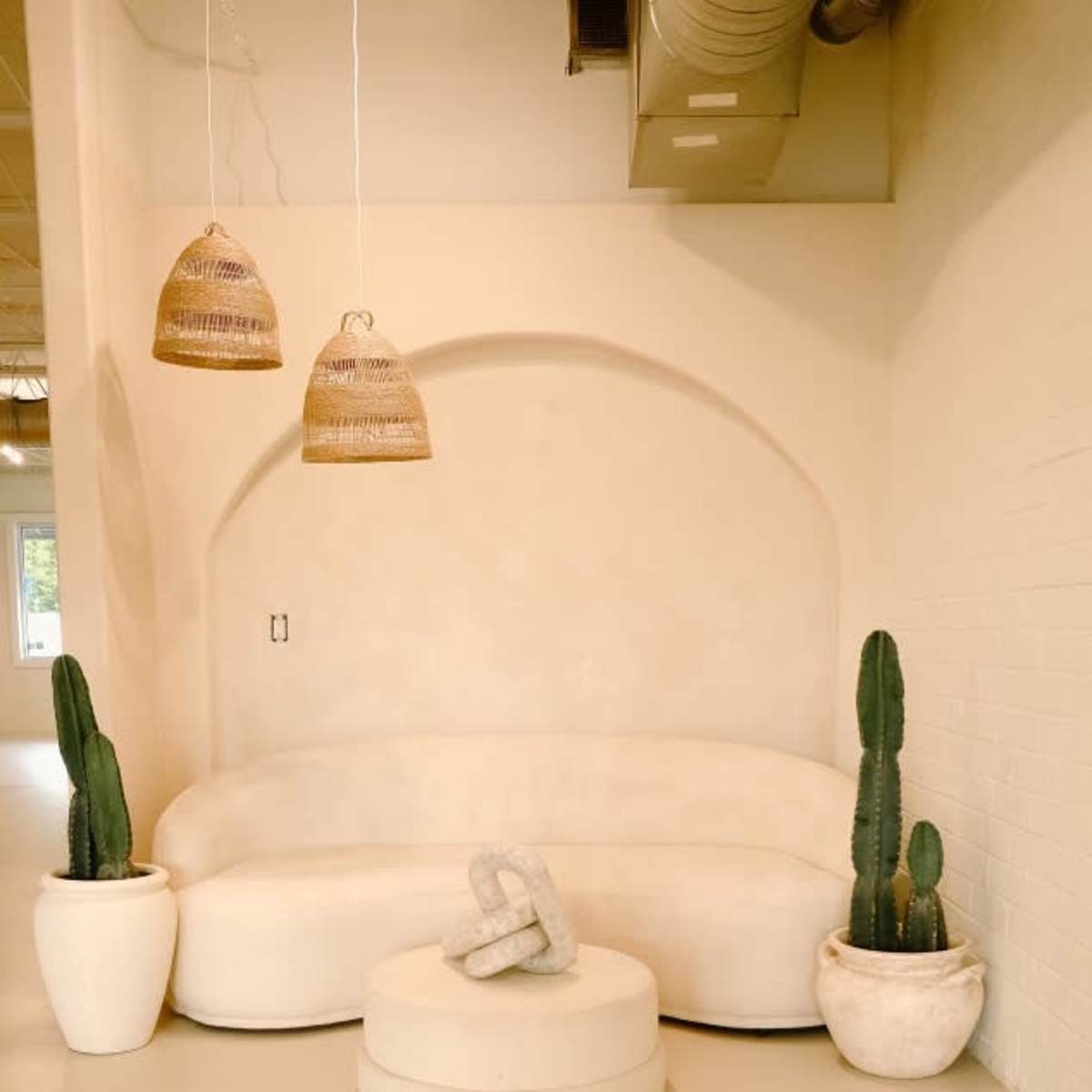 A minimalist interior with a curved white sofa, a round coffee table, and two potted cacti, complemented by two hanging woven light fixtures.