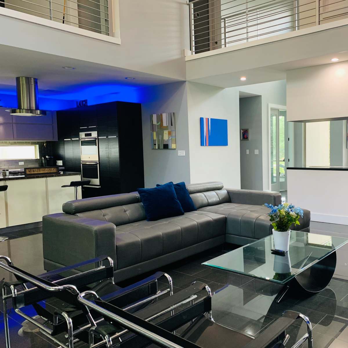 A modern living room with a gray sectional sofa, a glass coffee table, and an open layout leading to a contemporary kitchen with blue lighting.