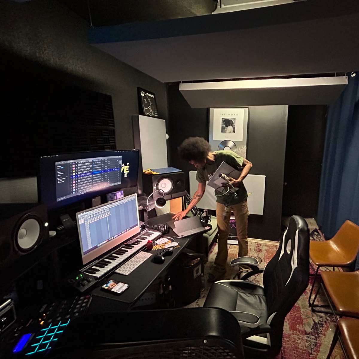 A person is arranging equipment in a music studio featuring a keyboard, monitors, and a computer setup.