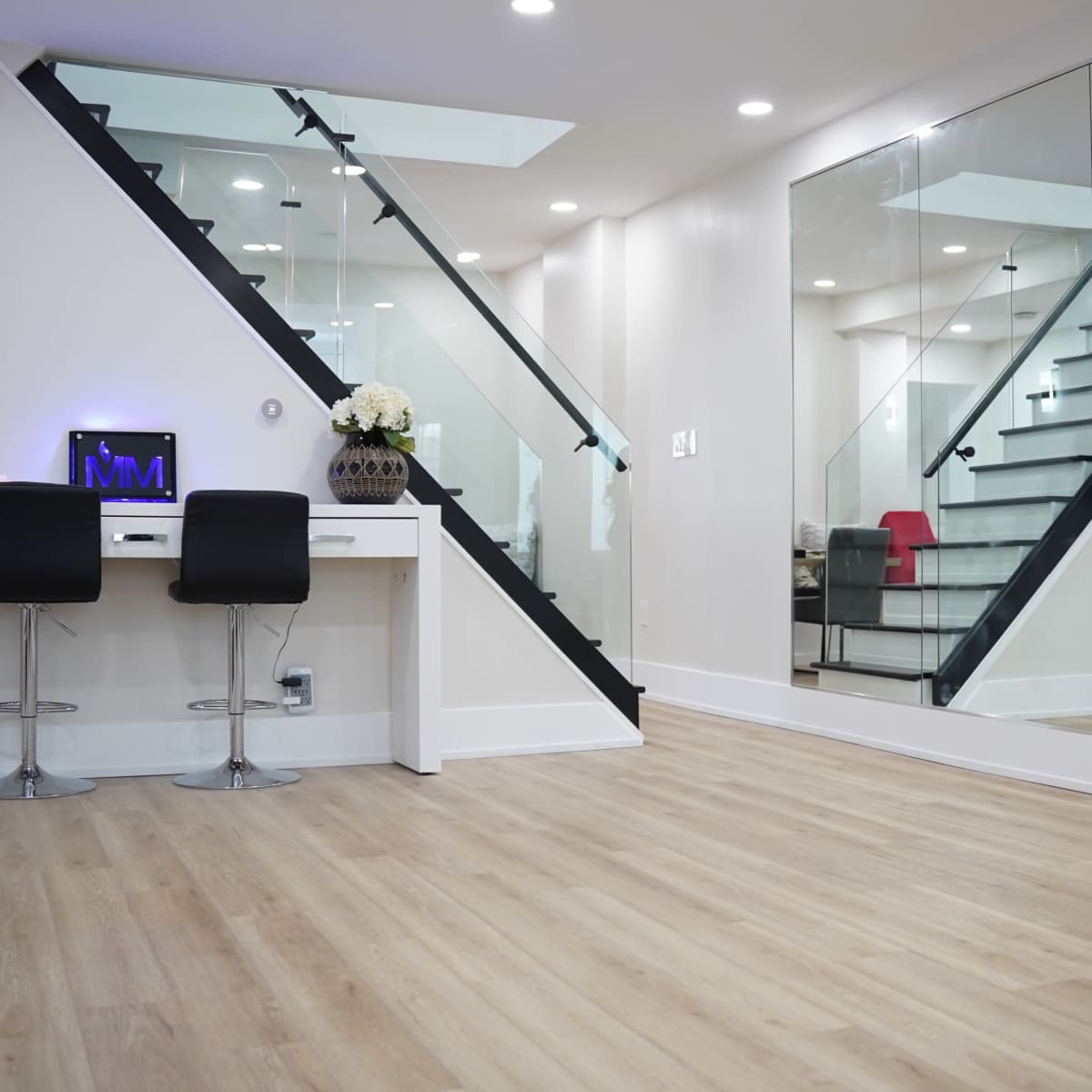 A modern interior space featuring a staircase with glass panels, a desk with two black chairs, and large mirrors reflecting the surroundings.