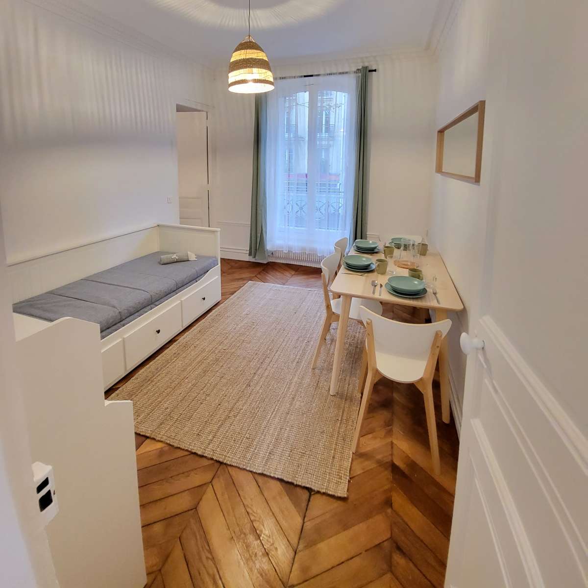 A brightly lit, minimalistic room featuring a bed, a dining table set for two, and a large window with light curtains.