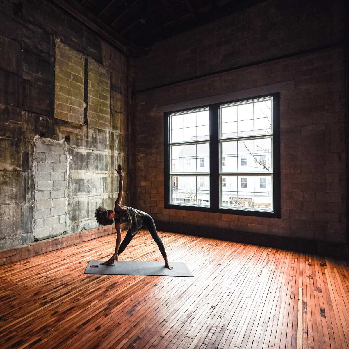 A person is performing a yoga pose on a mat in a room with large windows and wooden flooring.