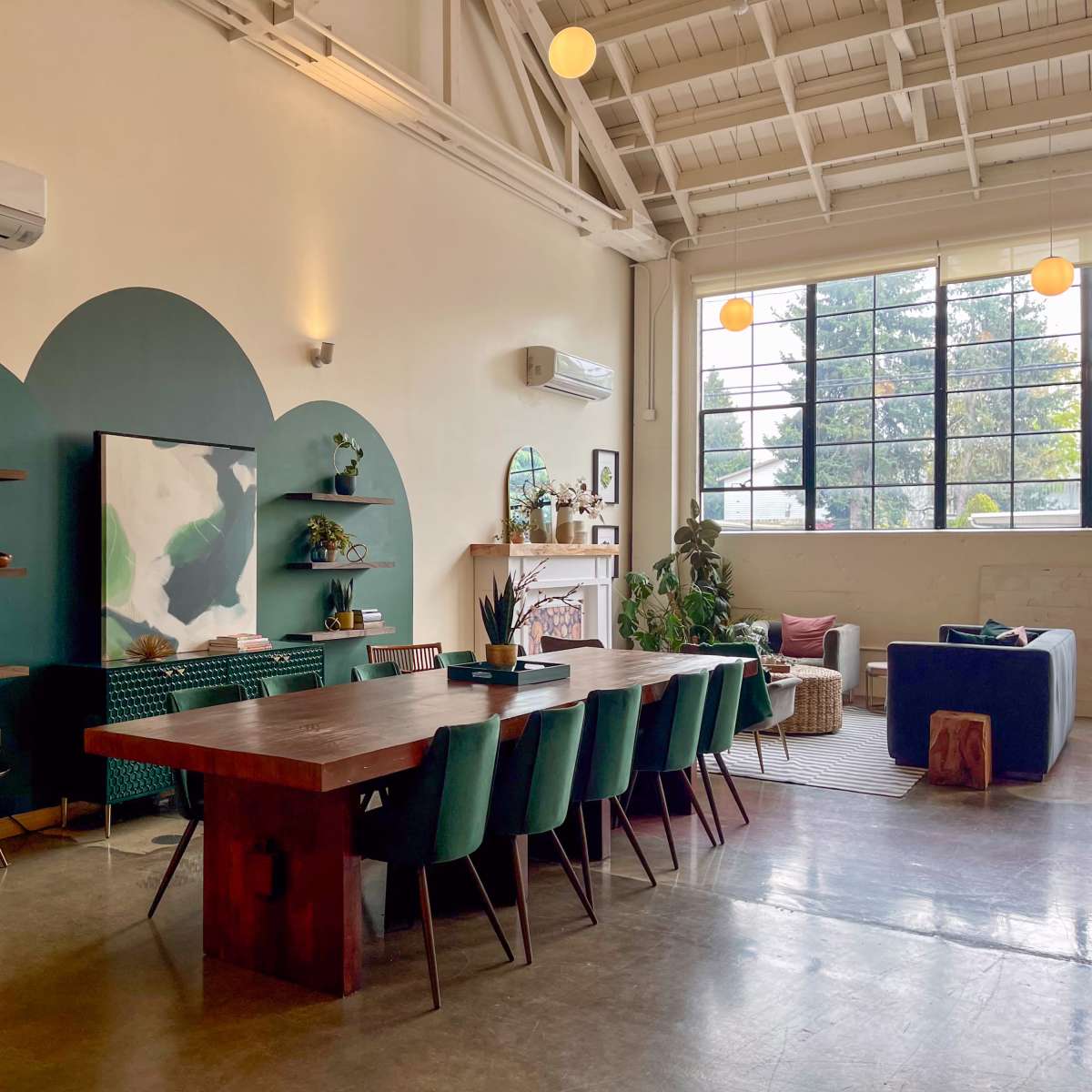 A spacious interior featuring a long wooden dining table with green chairs, a large wall-mounted TV, and various decorative items, all under a high ceiling with large windows allowing natural light.