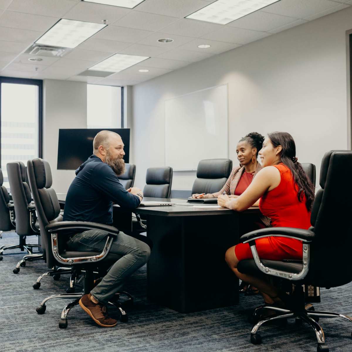 Three professionals are seated at a conference table in a modern office, discussing matters with a laptop in front of them.