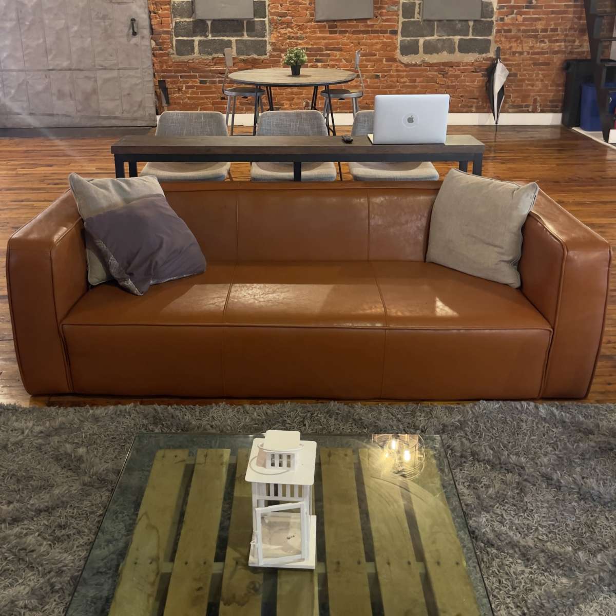 A brown leather sofa sits in the foreground of a room featuring exposed brick walls, a modern dining table with chairs in the background, and a laptop on the table.