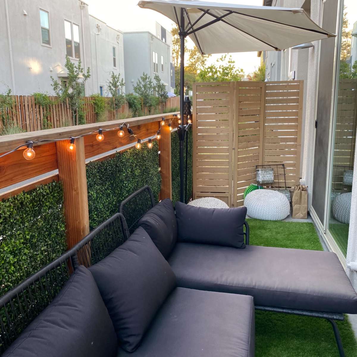 A small outdoor patio area with a black sofa, an umbrella, and wooden fencing, surrounded by grass and decorative lighting.