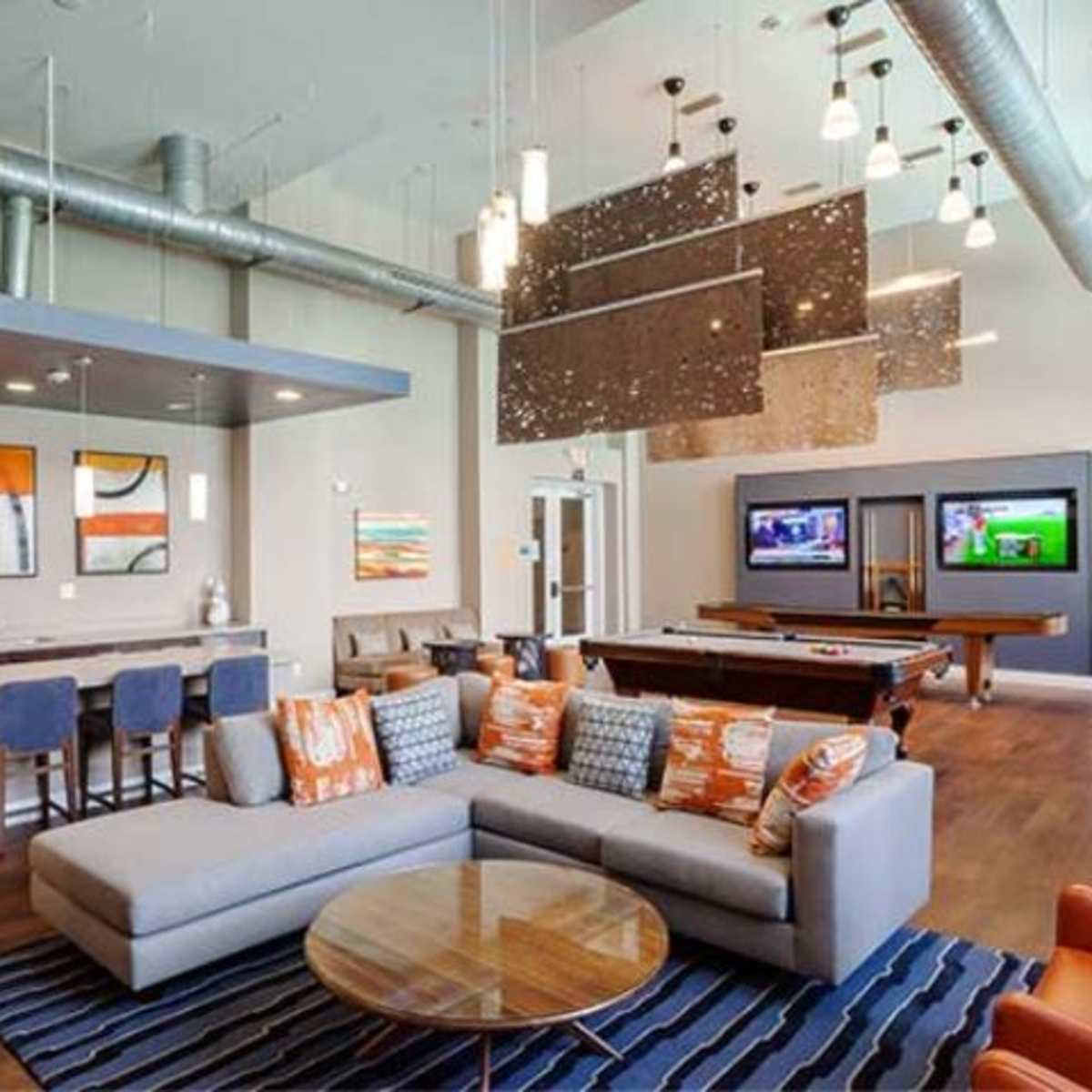 A modern lounge area featuring a sectional sofa, a coffee table, a kitchen bar with stools, and two wall-mounted televisions.