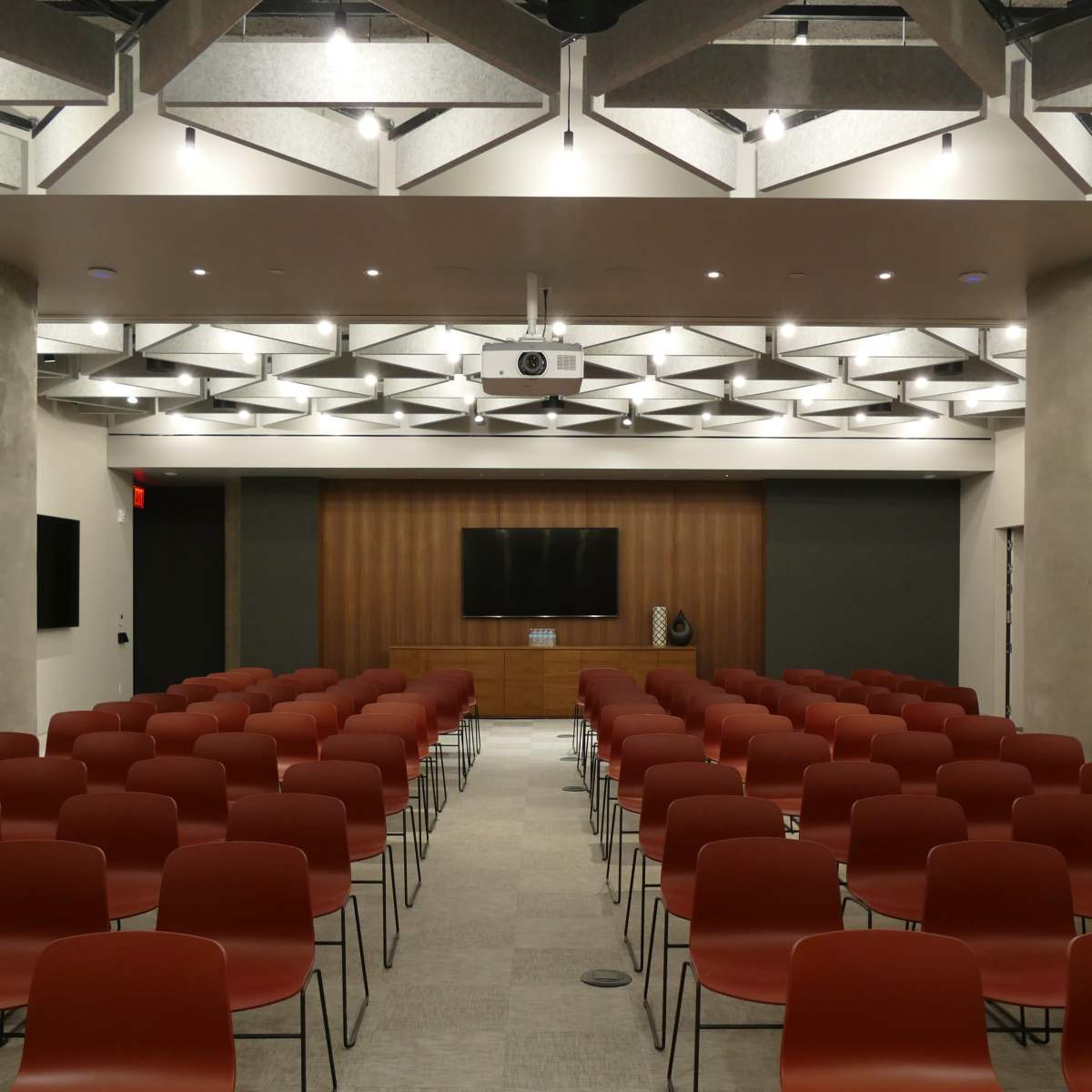 A conference room is set up with rows of red chairs facing a wooden panel at the front, featuring a large screen and a projector.