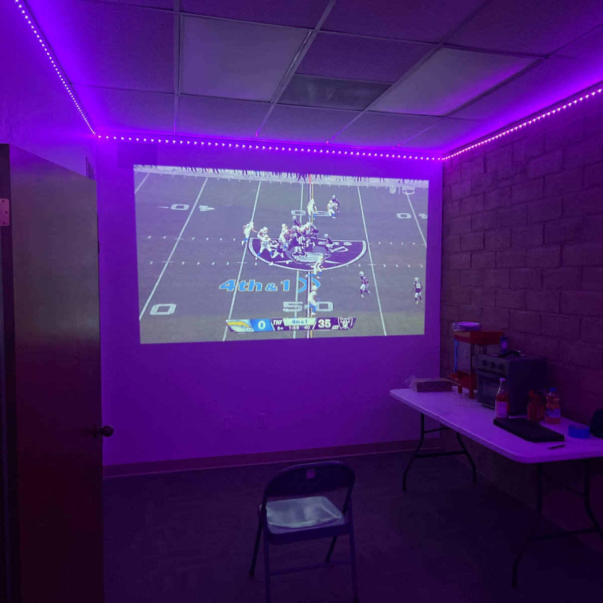 A football game is projected on a wall in a dimly lit room with purple LED lights.