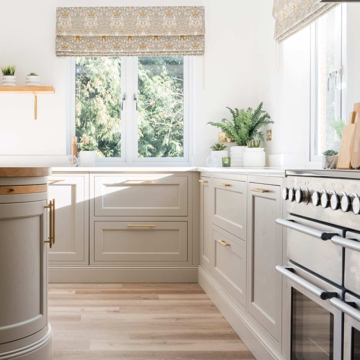 A well-lit kitchen features light-colored cabinetry, a wooden countertop, and plants arranged on shelves and windowsills.