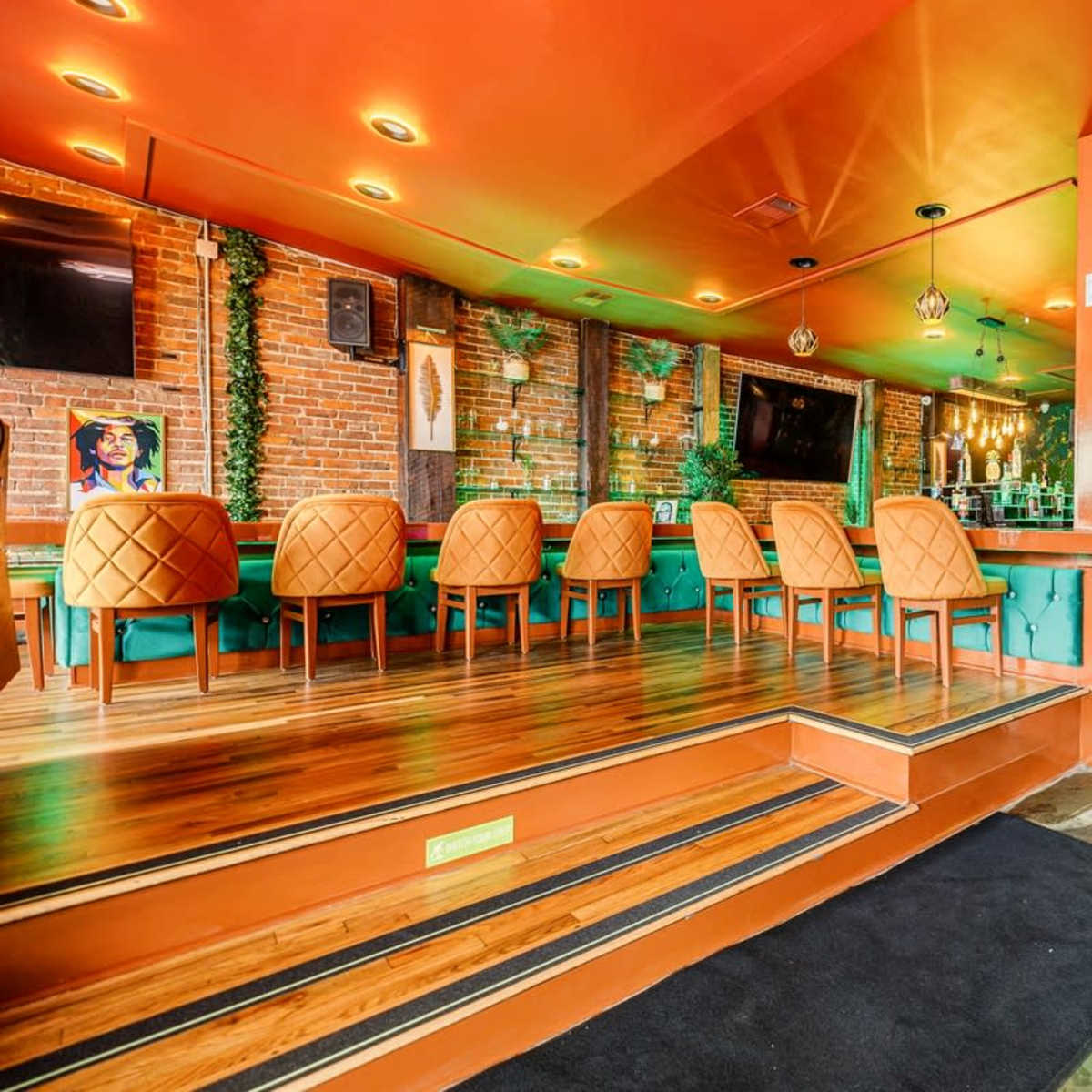 A vibrant bar interior with orange walls, green seating, and wooden flooring, featuring a mix of high and low seating arrangements.