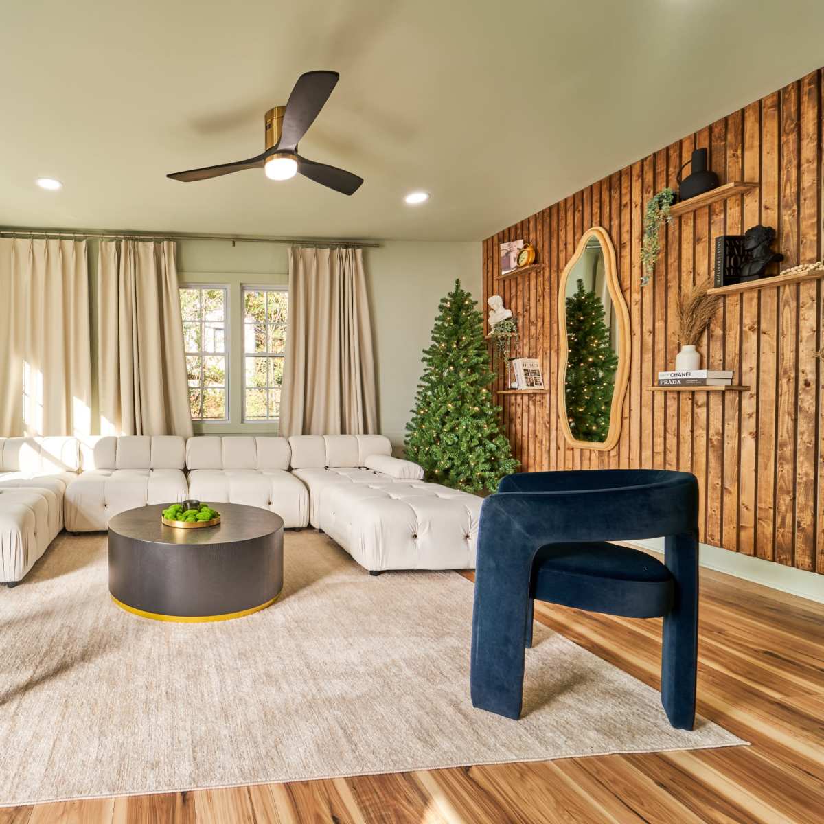 A modern living room features a white sectional sofa, two dark blue chairs, a round black coffee table, and floor-to-ceiling curtains, all set against a wooden accent wall adorned with decorative items.