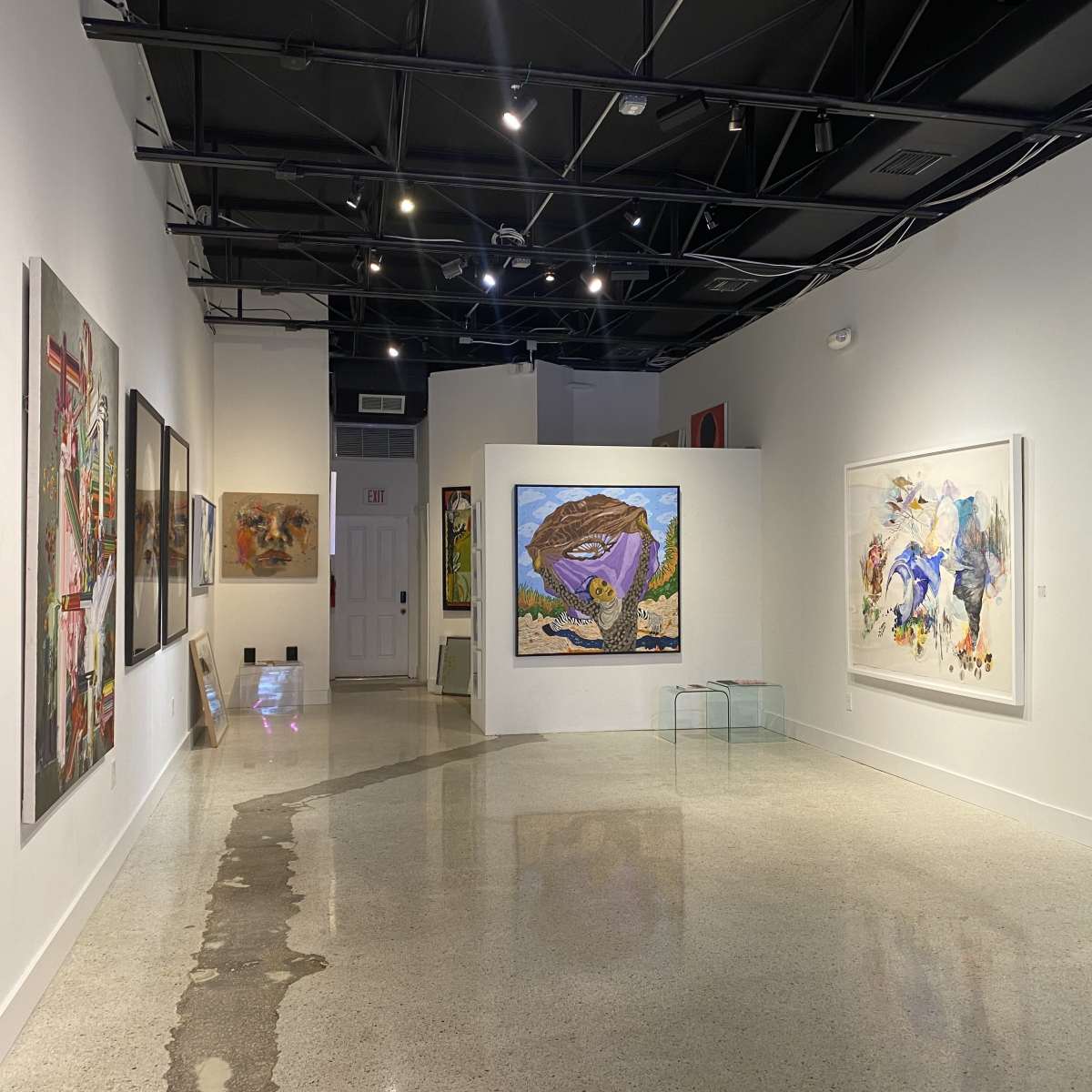 A well-lit art gallery featuring several paintings displayed on the white walls, with polished concrete flooring reflecting the light.