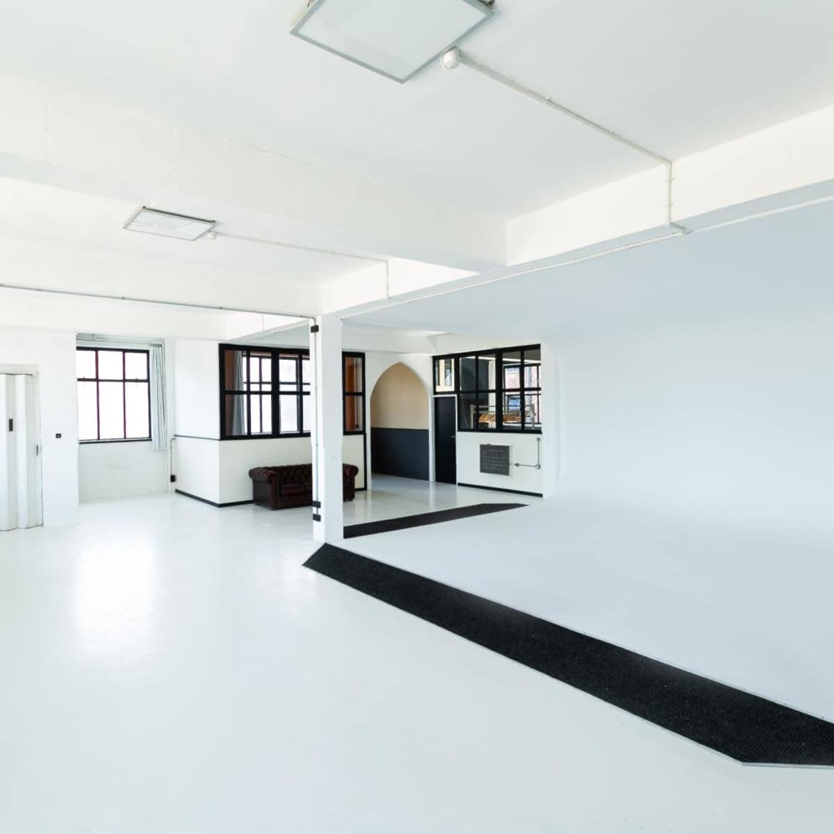A spacious, empty interior with white walls, a polished floor, and large windows, featuring a small partitioned area in the center.