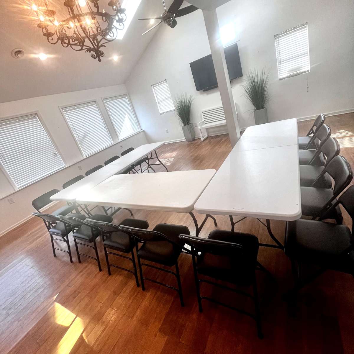 A large room features a setup of tables arranged in a U-shape with chairs, illuminated by a chandelier and natural light from windows.