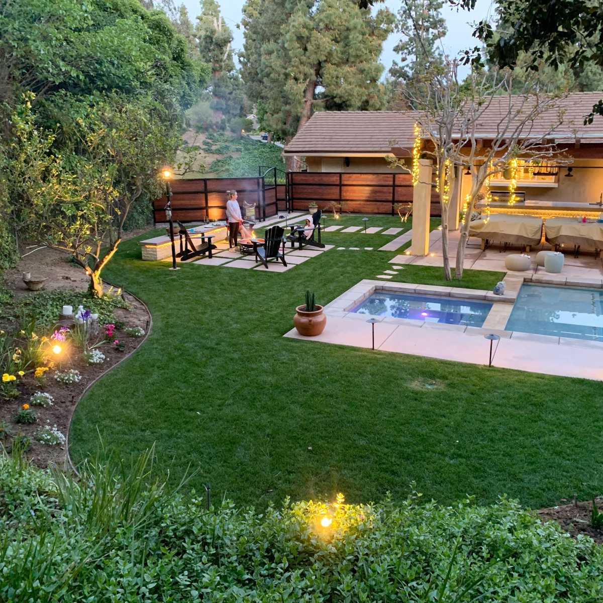 A landscaped backyard features a seating area around a fire pit, a swimming pool, and illuminated pathways amidst greenery and blooming flowers.