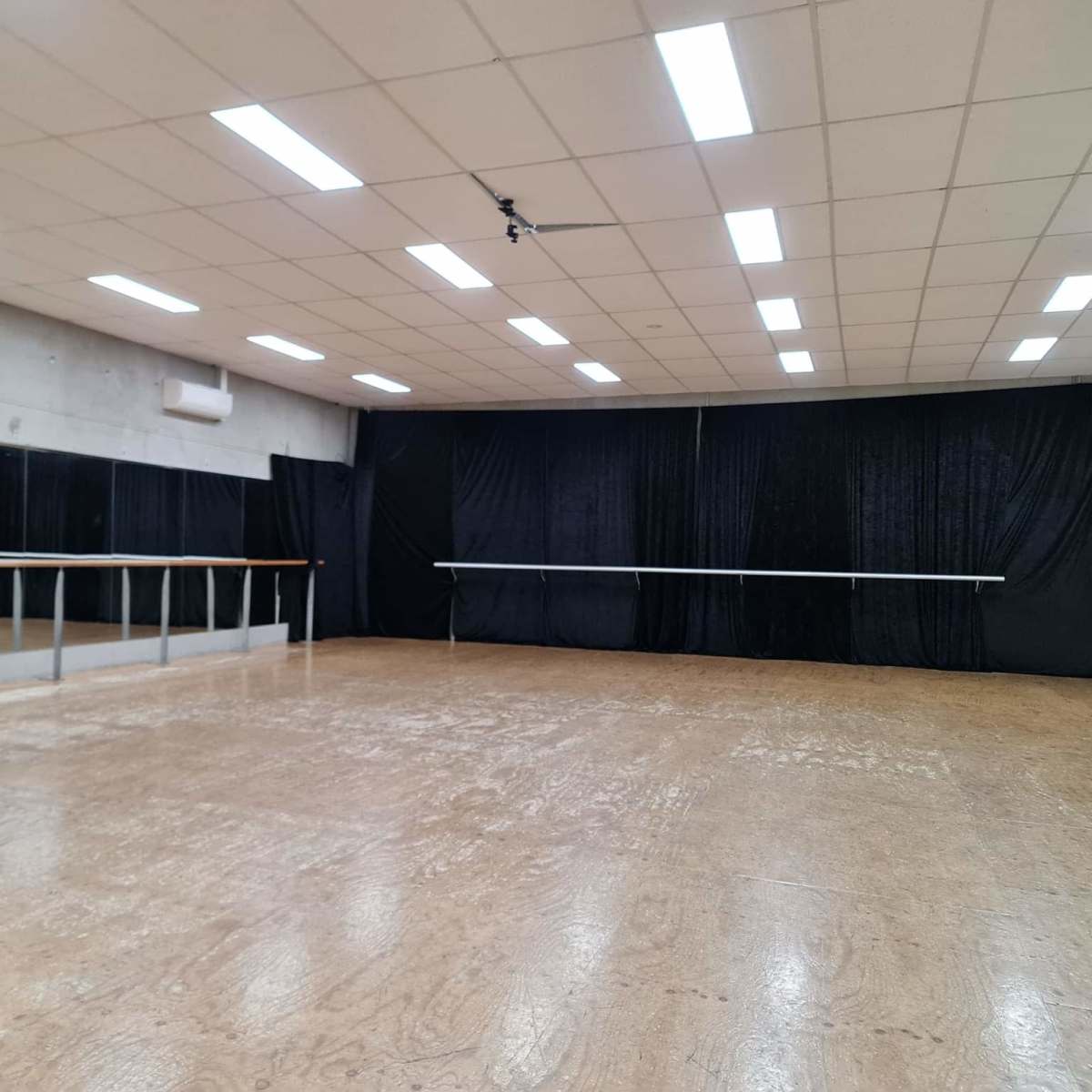 A spacious dance studio with a wooden floor, mirrored walls, and a black curtain at the far end.