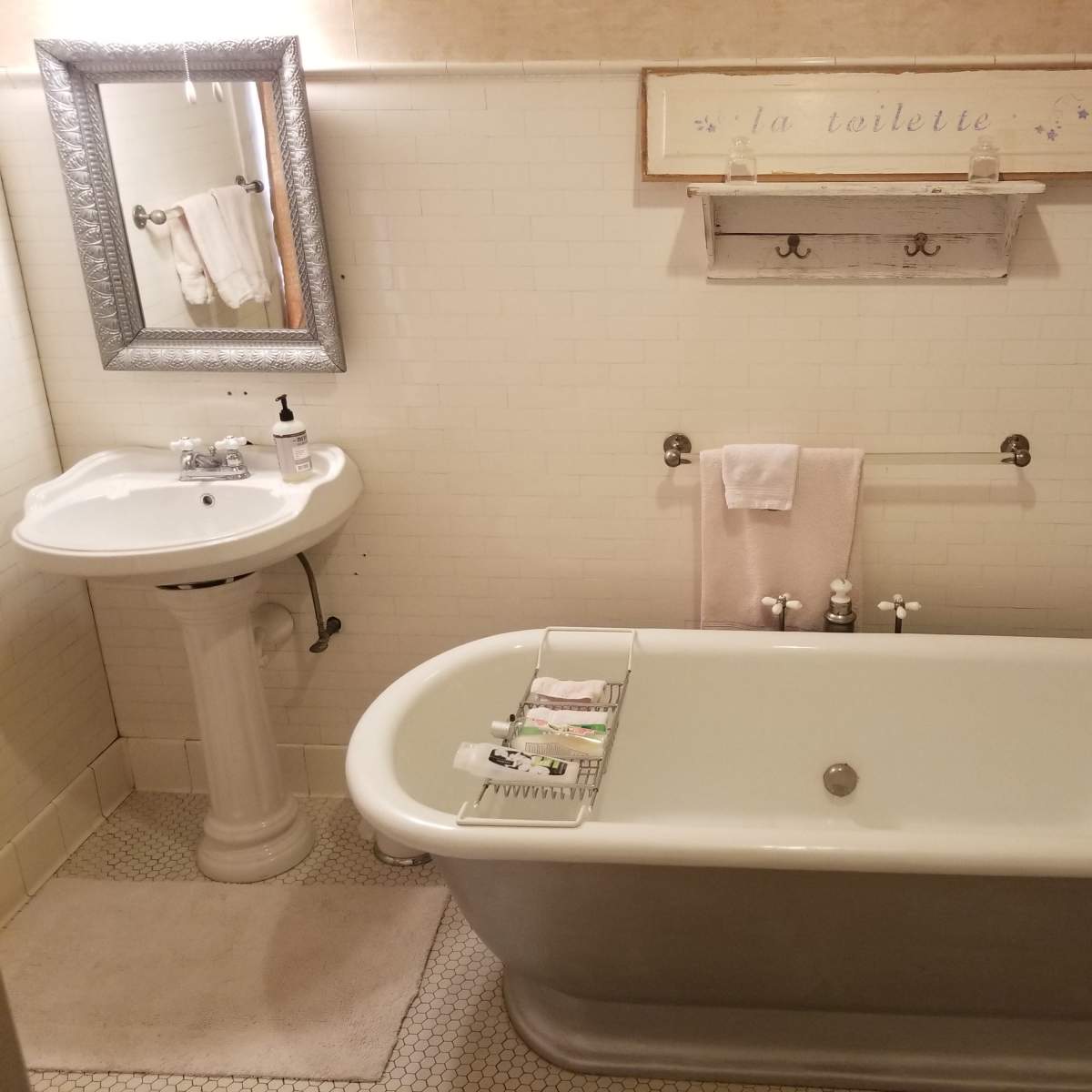 The bathroom features a clawfoot tub, pedestal sink, and a toilet, all arranged against tiled walls with a small shelf and decorative sign above.