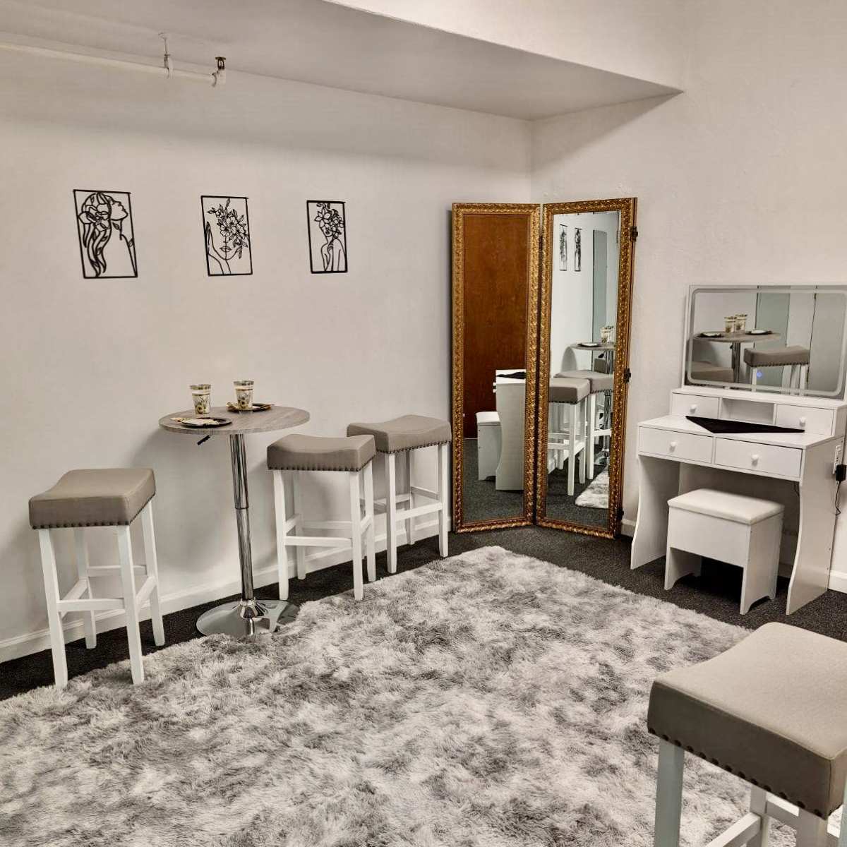 A minimalist room with a plush gray carpet, white furniture including stools and a vanity, and three framed artworks on the walls.