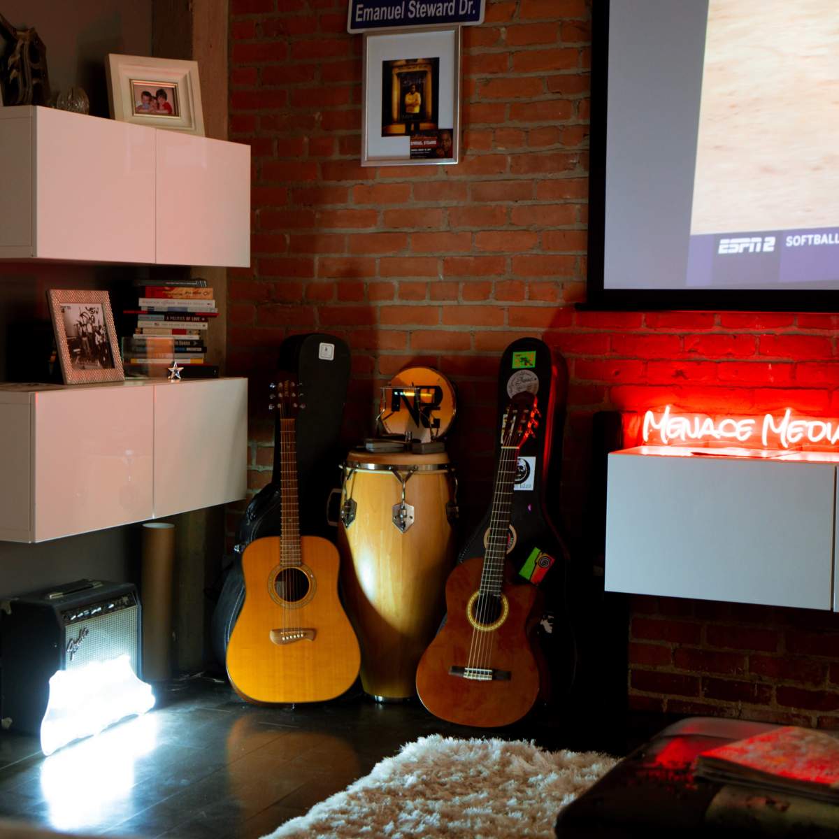 A cozy living room corner with white wall-mounted shelves, a collection of guitars and a conga drum, and a glowing neon sign.