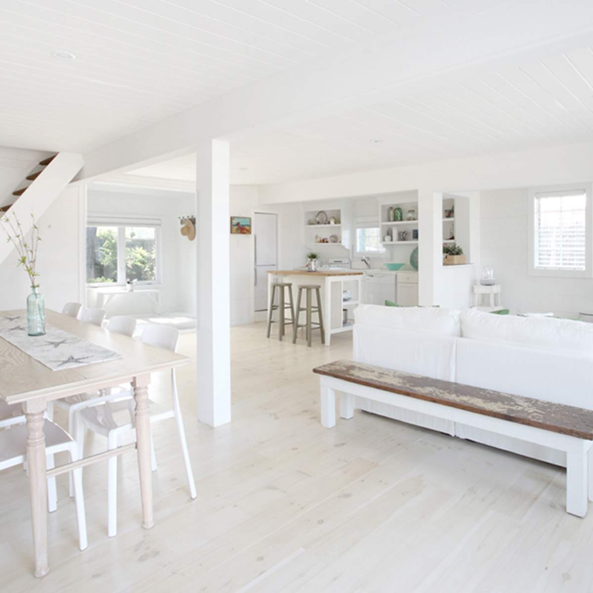A bright, open living space with a white color scheme, featuring a dining area with a wooden table and chairs, a sitting area with a white sofa, and a staircase in the background.
