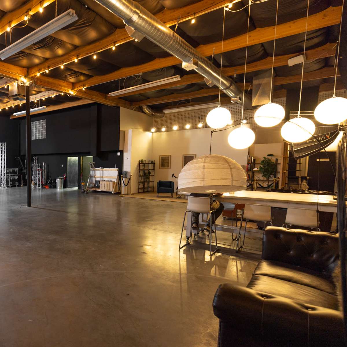 A spacious studio with high ceilings, concrete flooring, and various equipment, including a couch, lighting fixtures, and a staircase in the background.