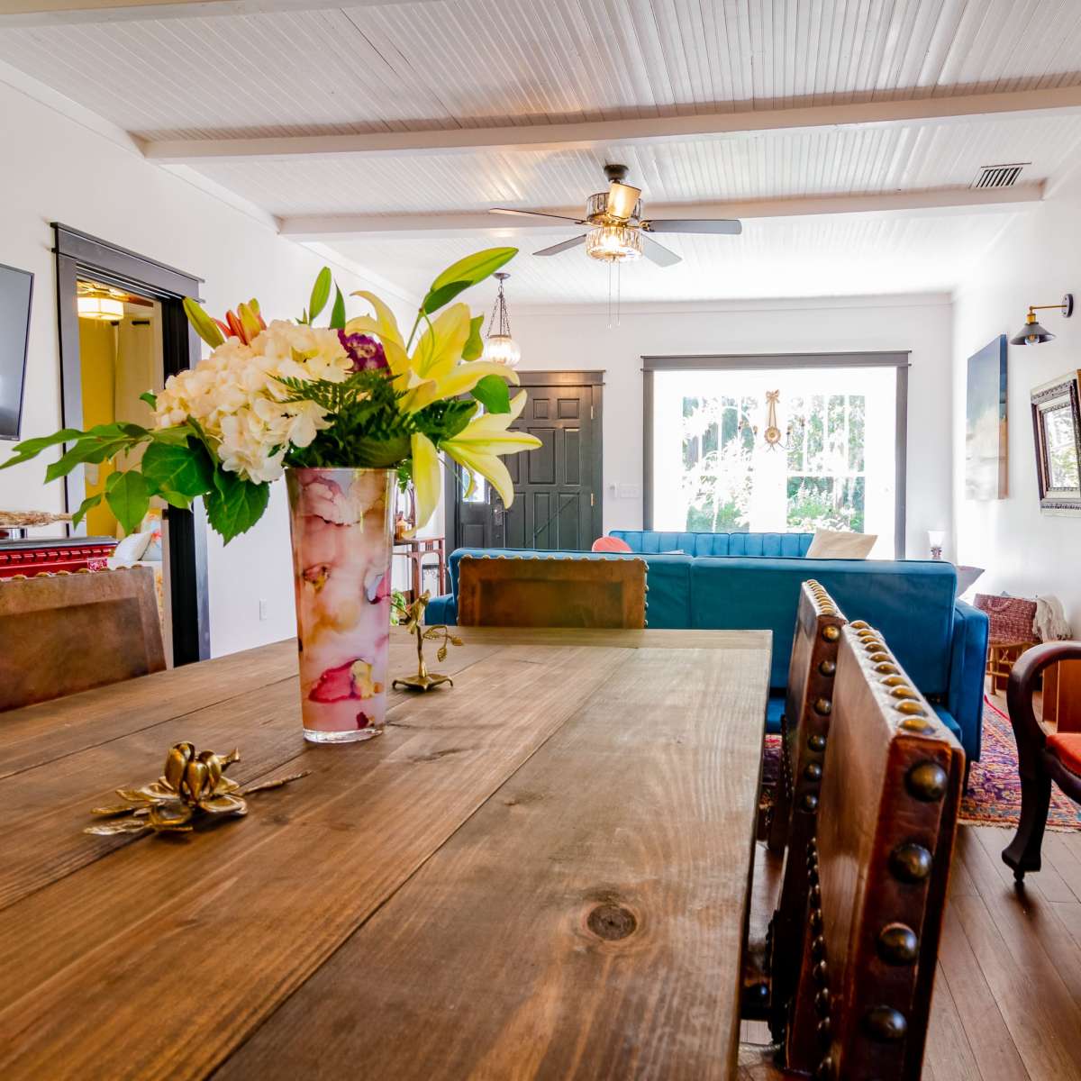 A dining area with a wooden table adorned with a floral arrangement, surrounded by chairs, and featuring a view of a living room with a blue couch and large windows.
