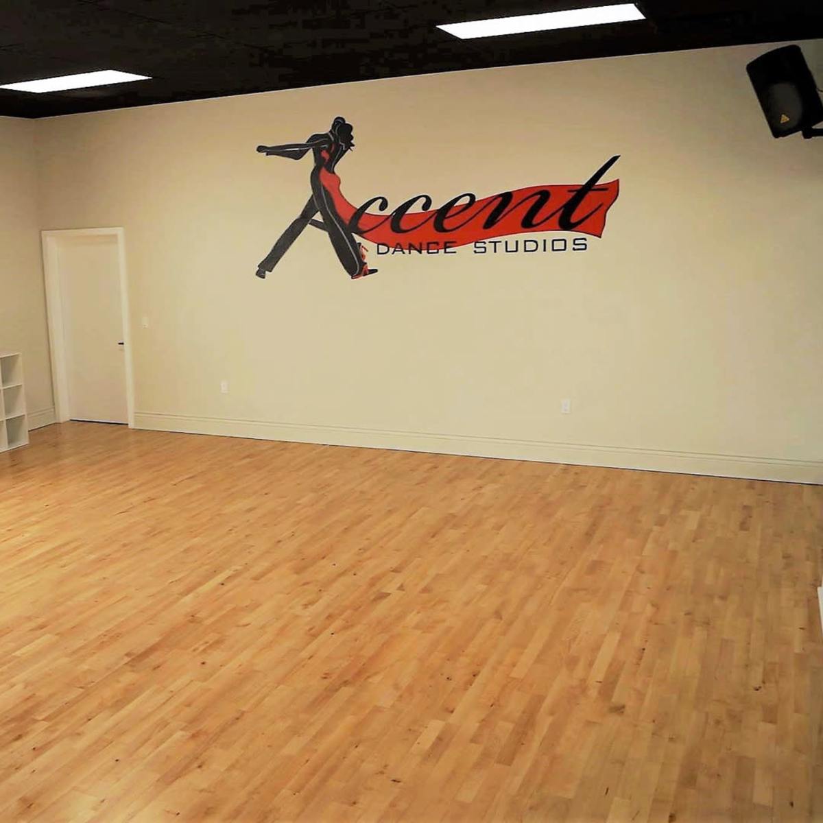 An empty dance studio with wooden floors, a large window, and a wall featuring a sign that reads "Accent Dance Studios."