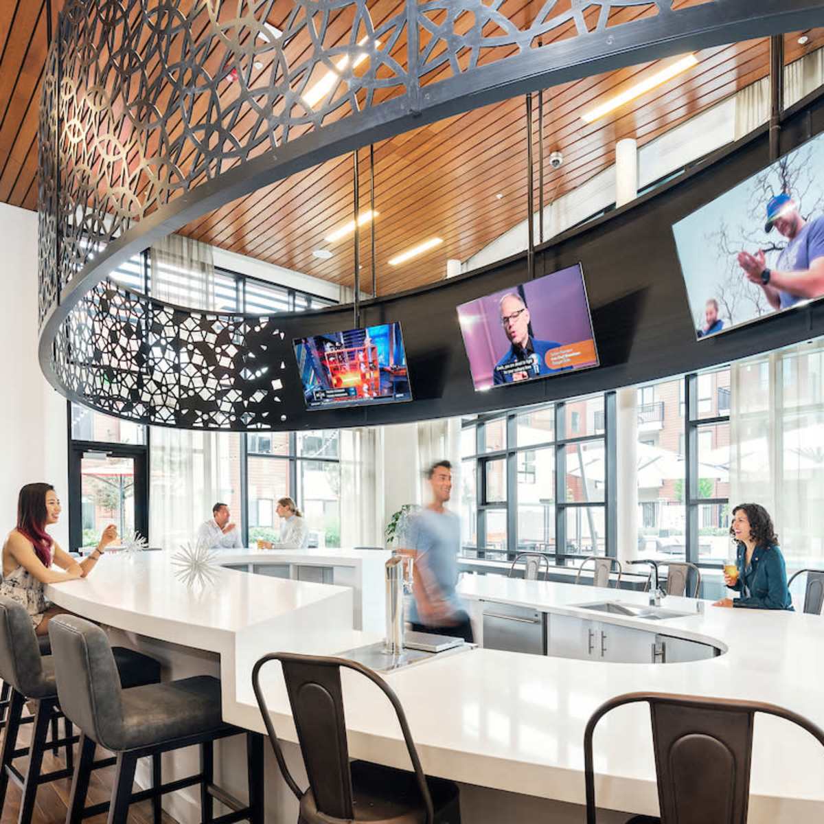 A modern communal space with a large white countertop, seating areas, and multiple televisions displaying various content.